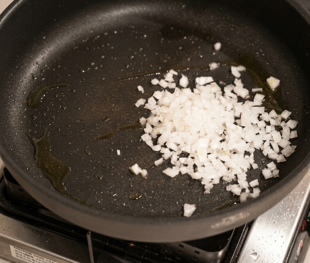 Cipolla bianca tritata che appassisce in una padella scura fino a diventare trasparente e morbida.