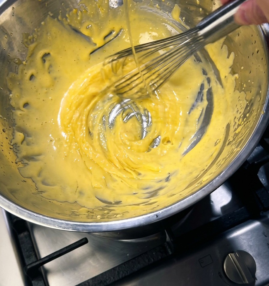 Una frusta metallica che mescola un composto denso e giallo chiaro in una ciotola dacciaio mentre viene versato un filo sottile di burro dorato.