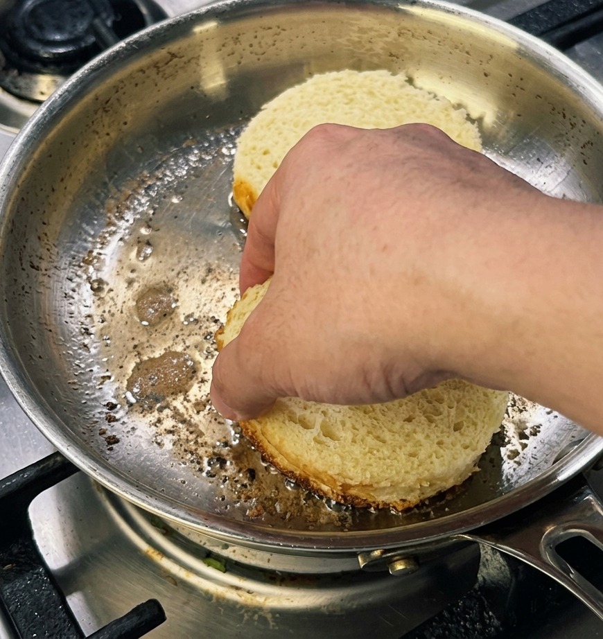 Una mano che preme due fette spesse e rotonde di pane brioche in una padella per ottenere una crosticina dorata.