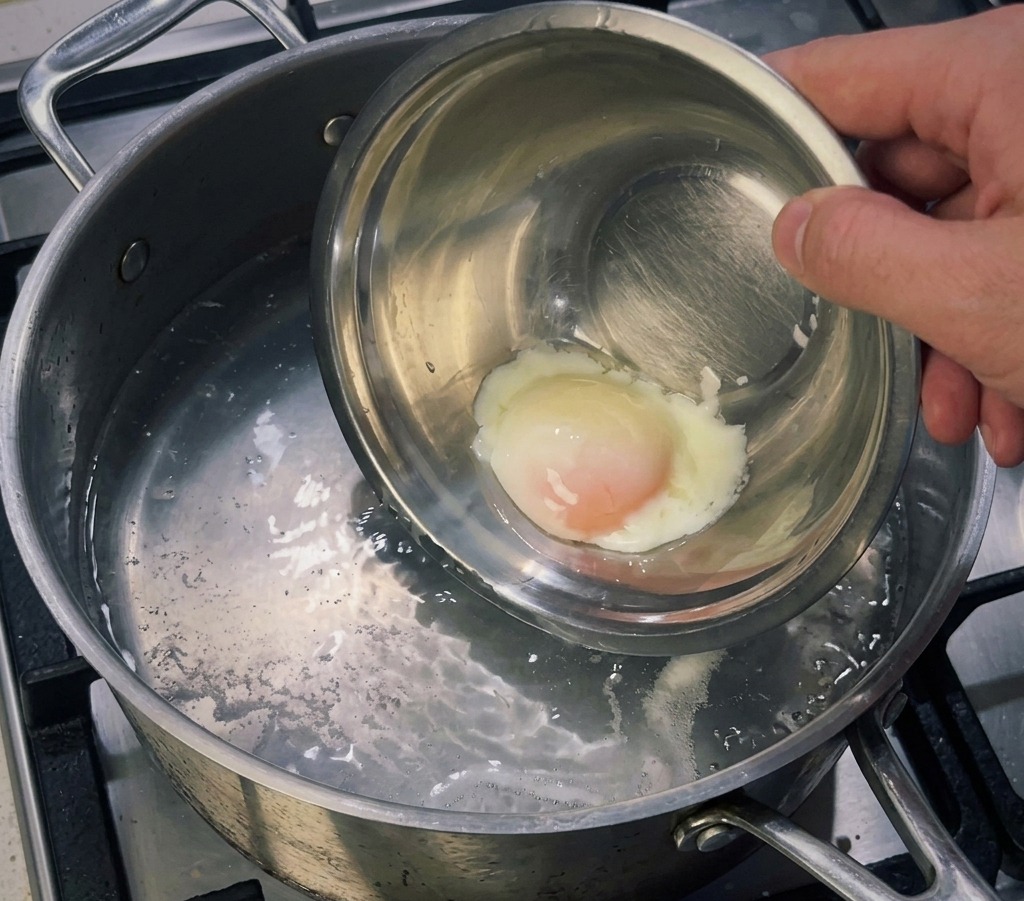 Una piccola ciotola di metallo con un uovo cotto a bassa temperatura che viene immerso in un pentolino di acqua bollente per rassodare lalbume esterno.
