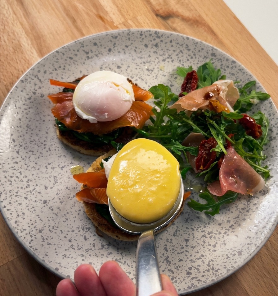 Primo piano di un grande cucchiaio che versa una salsa olandese densa, lucida e gialla su un uovo in camicia adagiato su salmone affumicato e brioche.