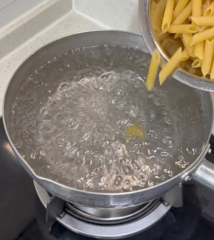 Penne rigate secche che vengono versate da una ciotola in acciaio inossidabile in una pentola dacqua bollente sul piano cottura.