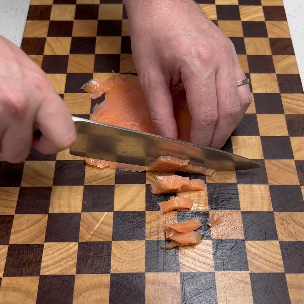 Mani di uno chef che usano un coltello dargento per tagliare fette piatte di salmone affumicato in piccoli quadratini su un tagliere a scacchi marrone e beige.
