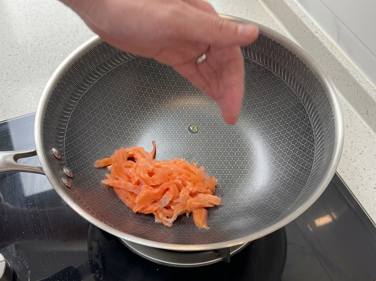 Fette di salmone affumicato rosa che vengono saltate in una padella in acciaio inossidabile con un motivo esagonale strutturato.