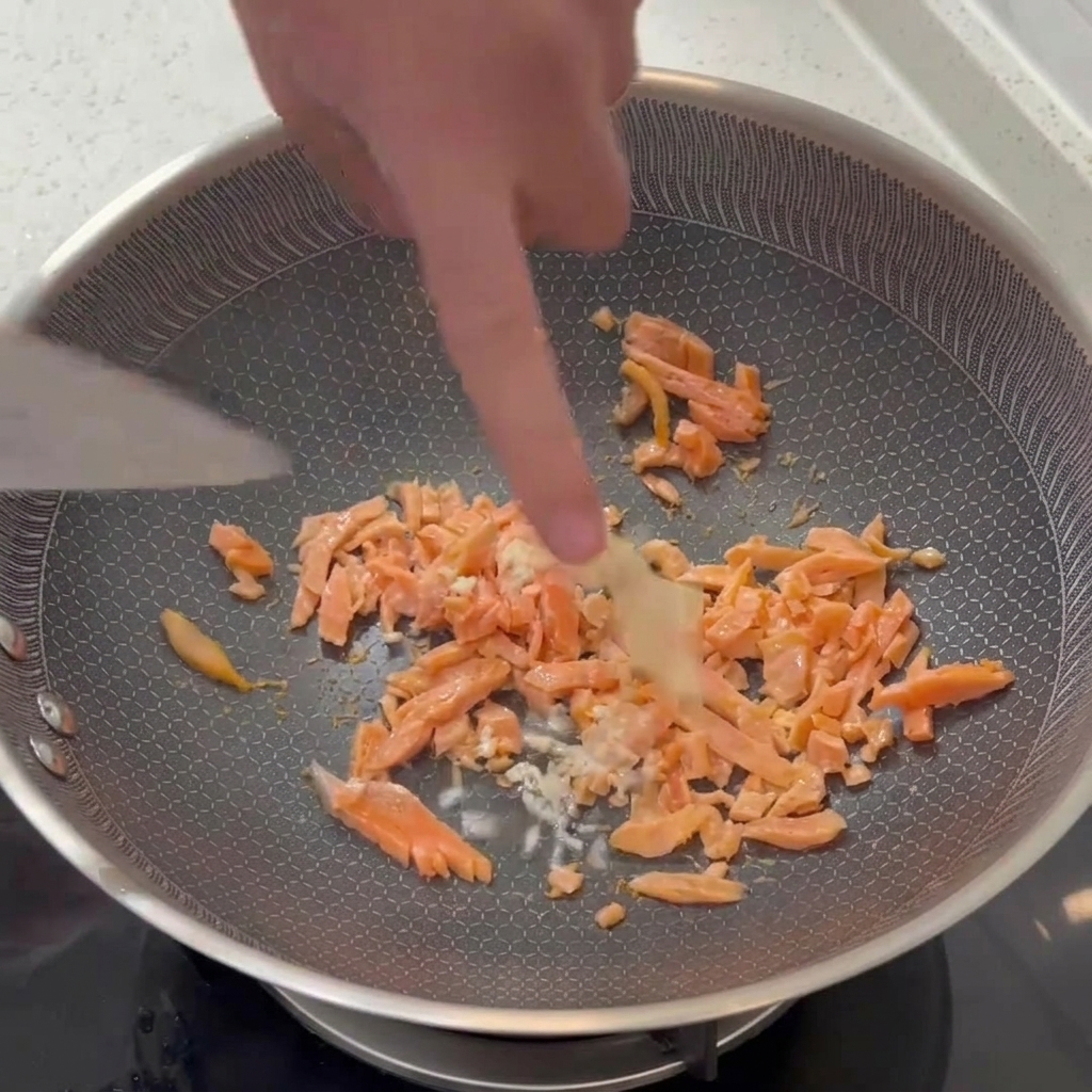 Aglio bianco tritato che viene mescolato con una spatola in una padella insieme a pezzetti di salmone affumicato parzialmente cotti.