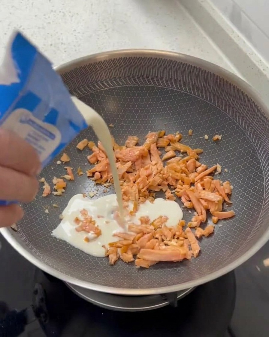 Una mano che versa la panna da cucina bianca da un brick blu e bianco in una padella contenente i pezzi di salmone cotti.