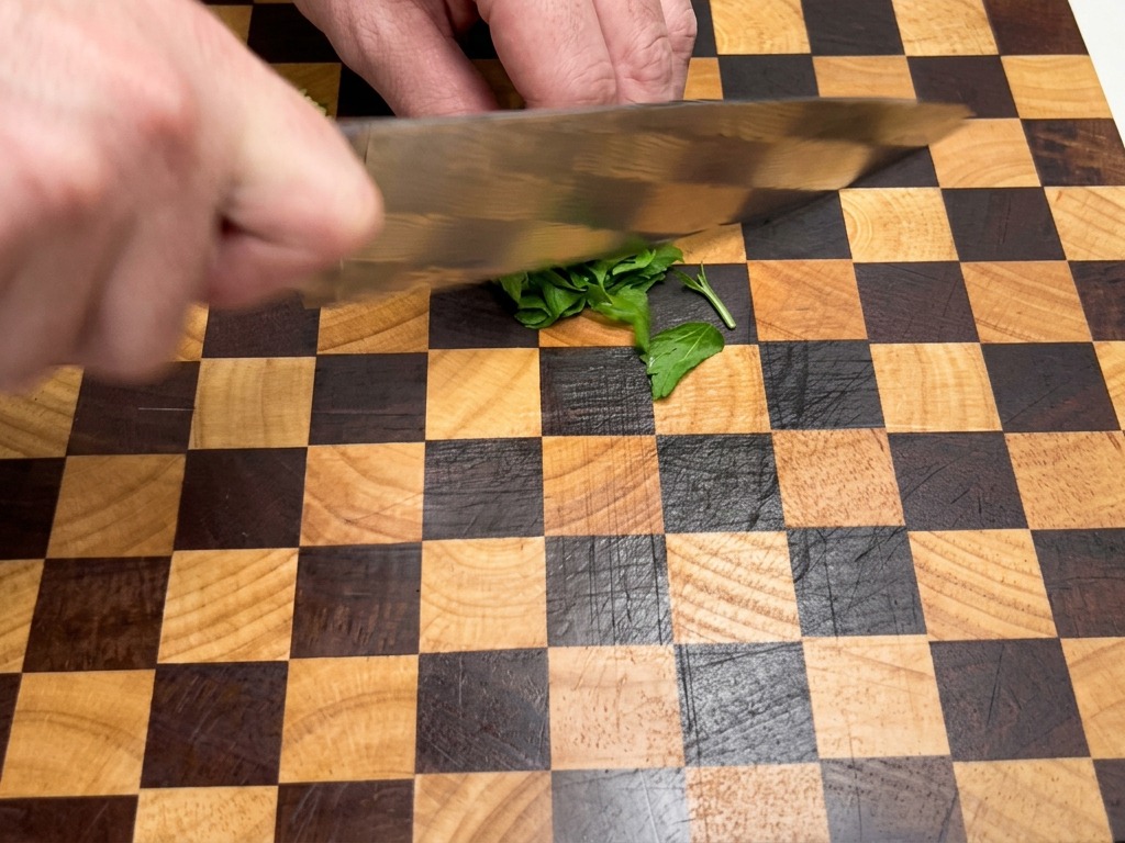 Primo piano di mani che usano un coltello da chef per tritare finemente foglie di prezzemolo verde fresco su un tagliere di legno a scacchi.
