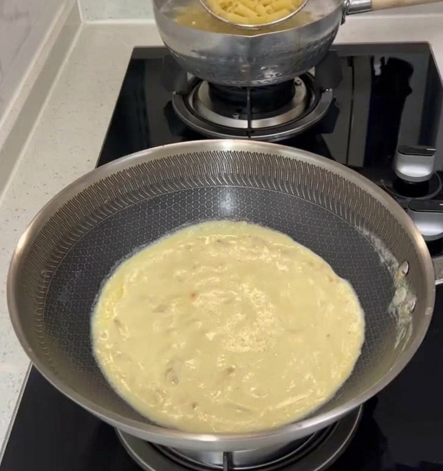 Un mestolo a ragno in metallo che raccoglie le penne cotte da una pentola bollente e le trasferisce in una padella piena di salsa alla panna cremosa sul piano cottura.