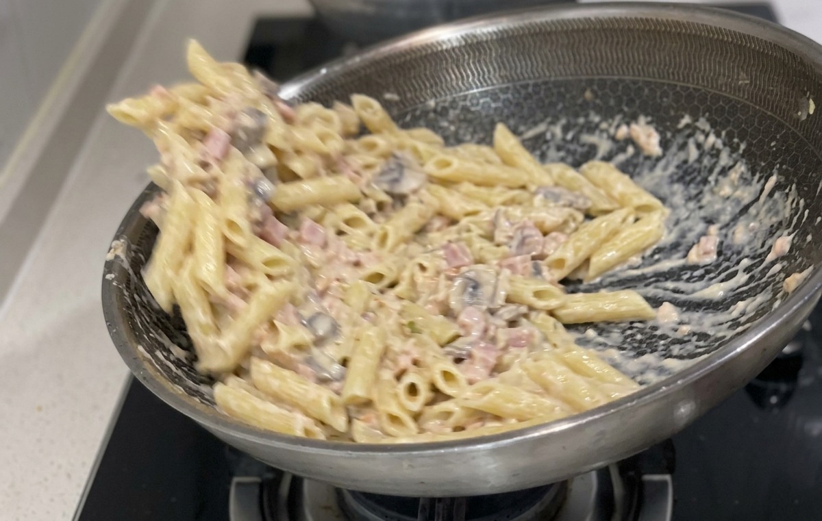 Penne cotte che vengono saltate e mescolate con una salsa bianca cremosa e pezzetti di salmone in una grande padella in acciaio inossidabile.