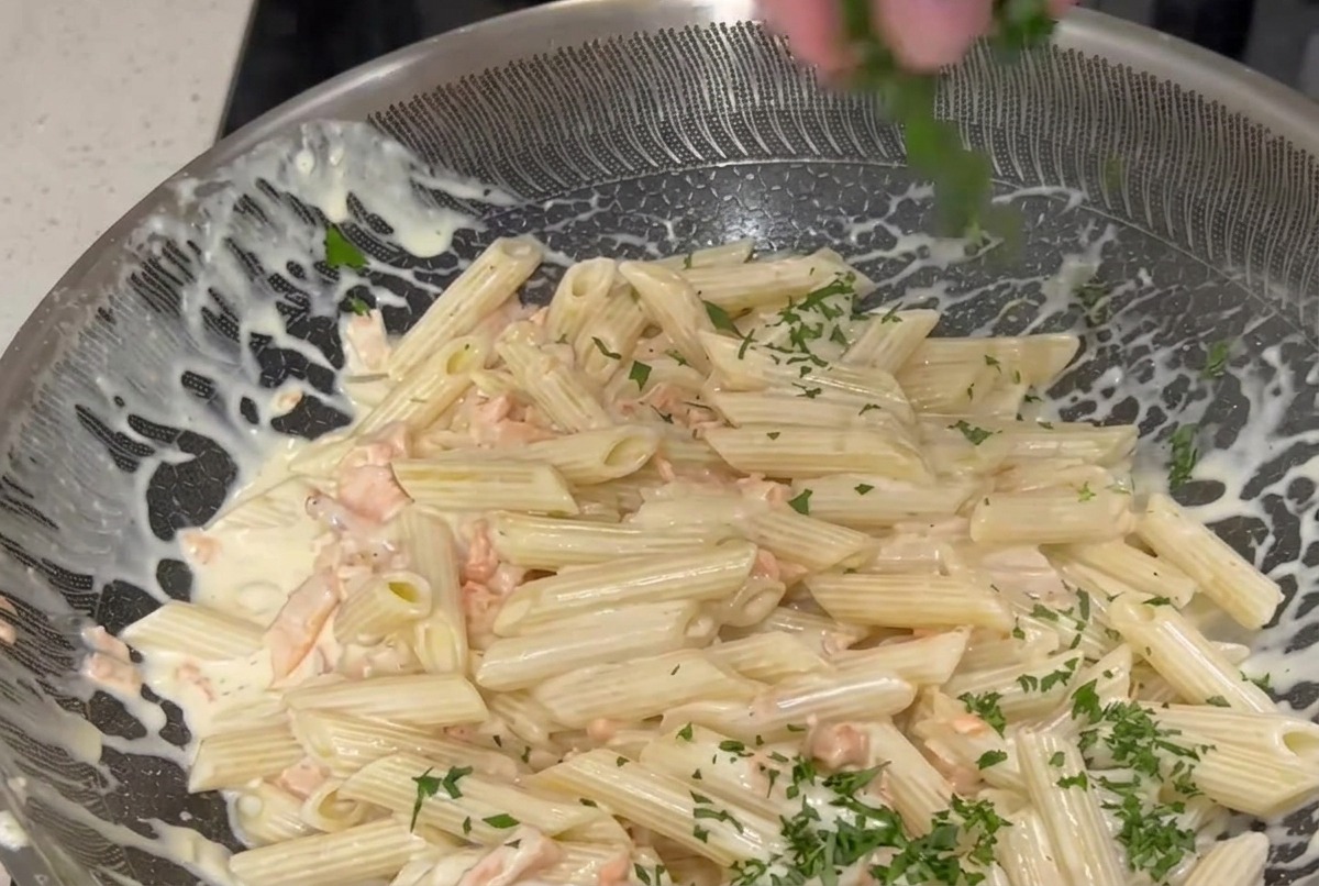 Una mano che distribuisce prezzemolo verde tritato finemente su una grande padella di penne condite con una salsa alla panna e pezzi di salmone affumicato rosa.