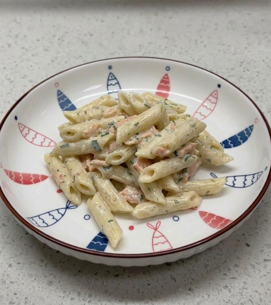 Una porzione di penne cremose al salmone elegantemente disposta in una ciotola di ceramica bianca decorata con un motivo di pesci blu e rossi lungo il bordo.