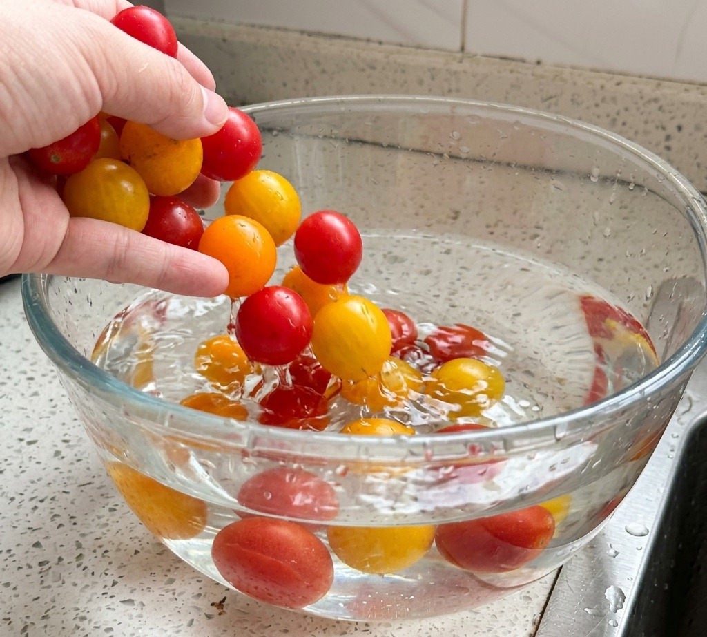 Mani che lavano pomodorini rossi e gialli in una ciotola di vetro piena dacqua.