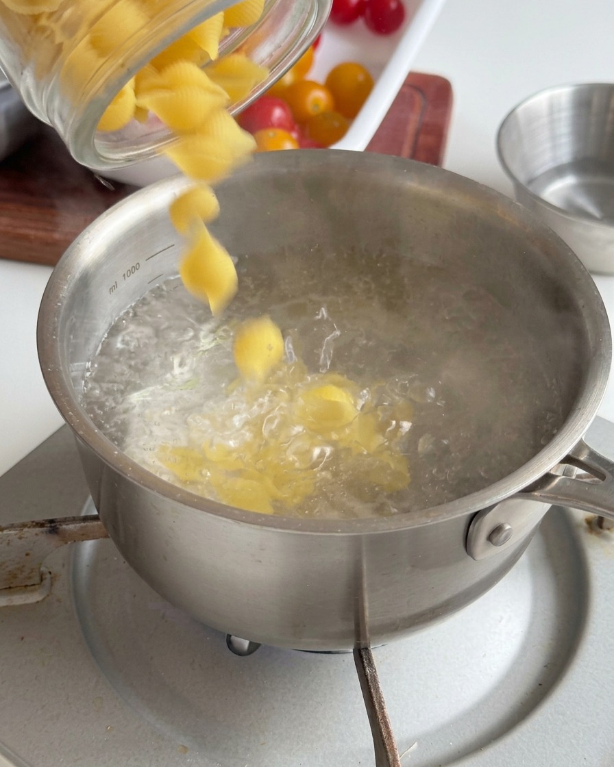 Conchiglie versate da un barattolo di vetro in una pentola dacciaio con acqua bollente.