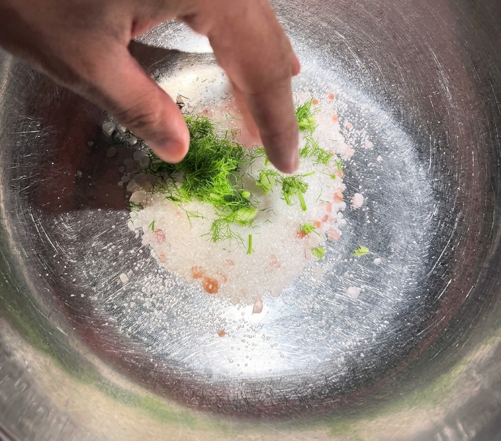 Una mano che mescola aneto verde fresco e spezie intere in un mucchio di sale bianco e zucchero allinterno di una ciotola di acciaio.
