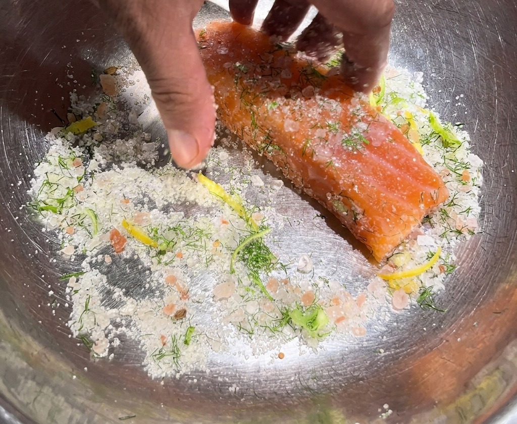 Mani che sfregano una miscela grossolana di sale, zucchero ed erbe su uno spesso pezzo di salmone crudo in una ciotola.