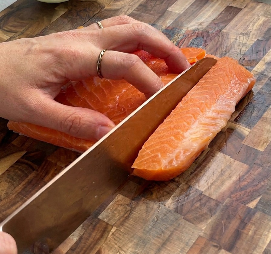 Primo piano di mani che usano un coltello per tagliare un filetto di salmone crudo a cubetti su un tagliere di legno.