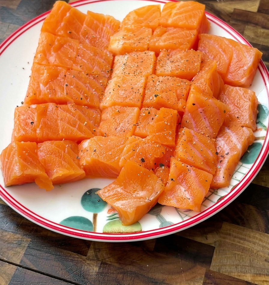 Cubetti di salmone su un piatto bianco, visibilmente conditi con grani di pepe nero e sale.