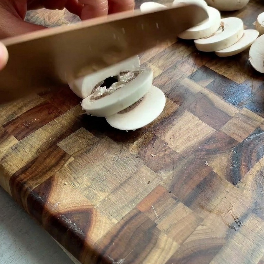 Mani che affettano funghi champignon bianchi freschi su un tagliere di legno.