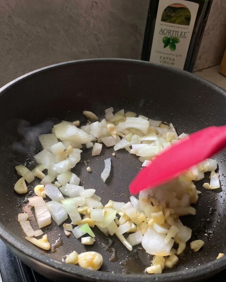 Una spatola rossa che mescola cipolla bianca tritata e aglio sminuzzato in una padella nera.