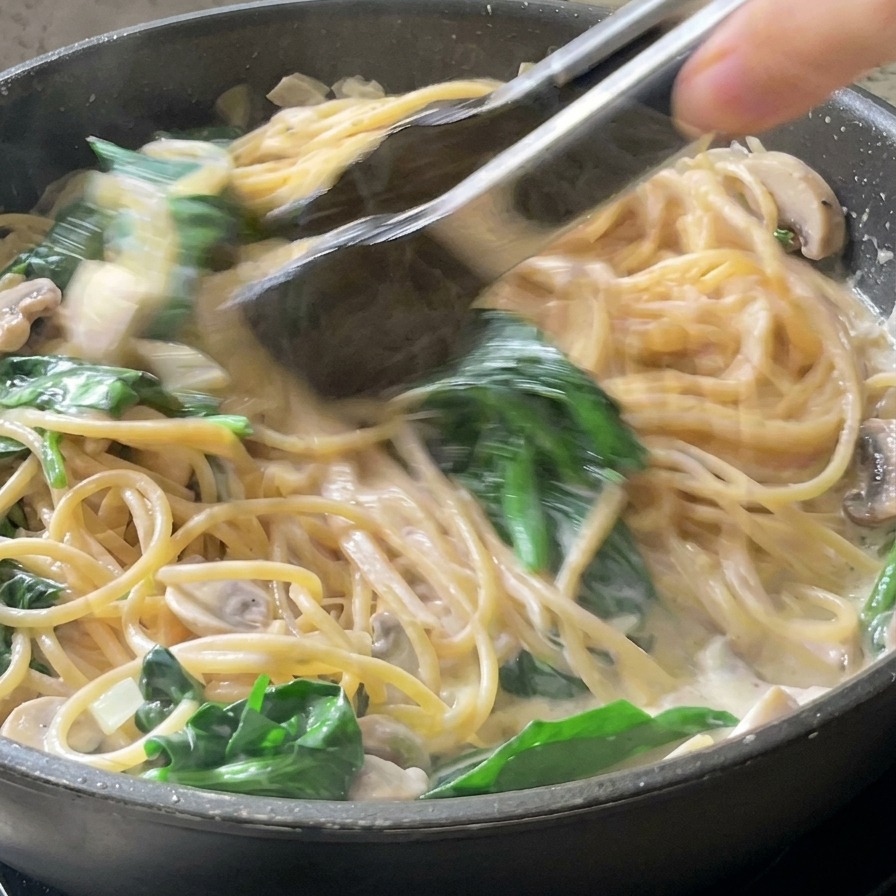 Pinze da cucina che tuffano spaghetti integrali in una salsa alla panna bianca con foglie di spinaci in una padella nera.