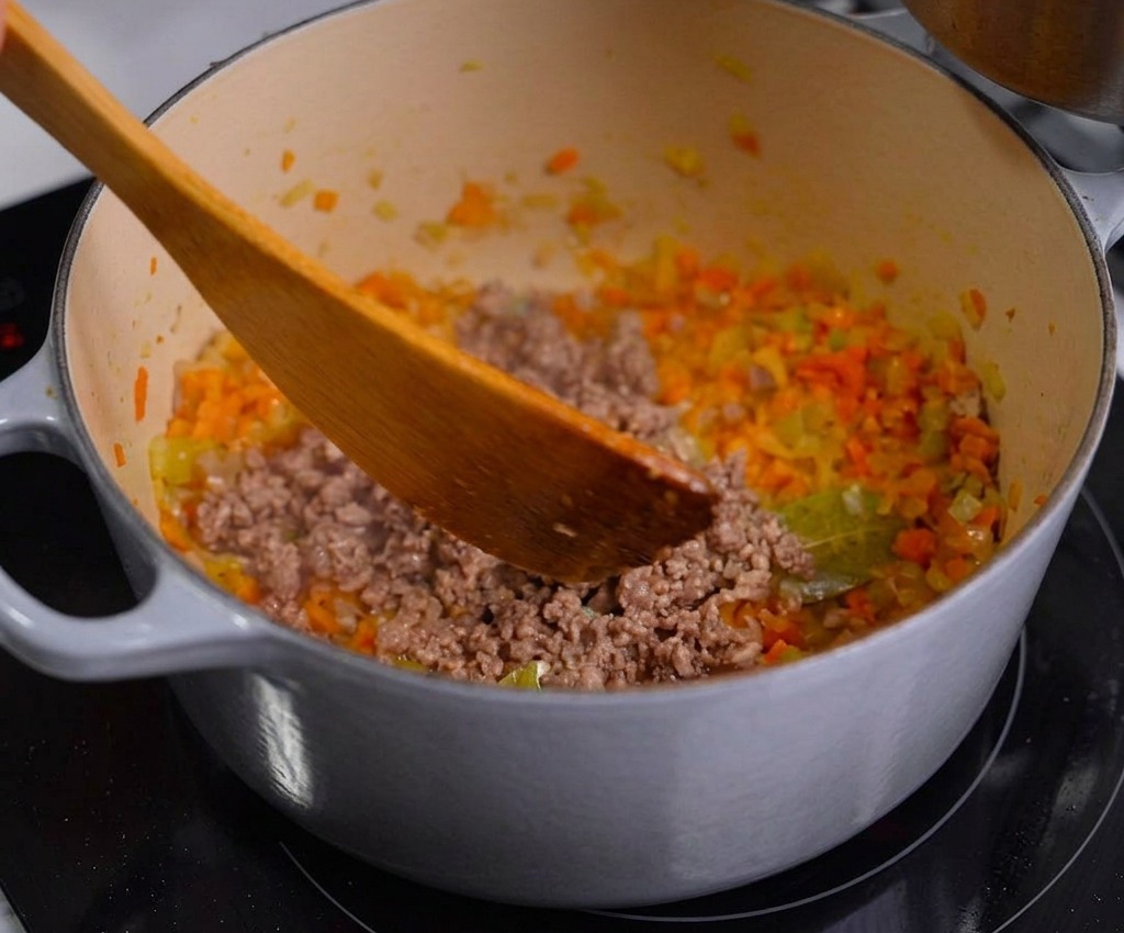 Una persona che usa una spatola di legno per incorporare la carne macinata rosolata nella pentola con le verdure soffritte.
