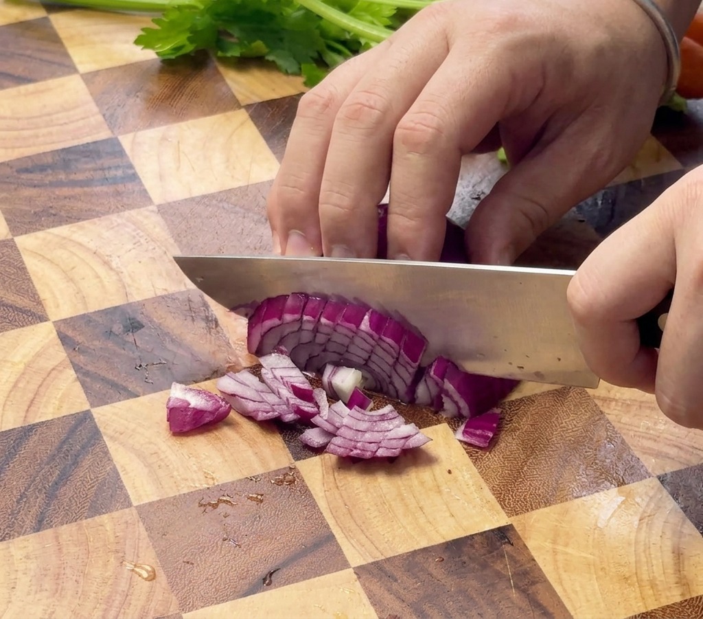 Primo piano di mani che tritano una cipolla rossa con un coltello da chef su un tagliere di legno.