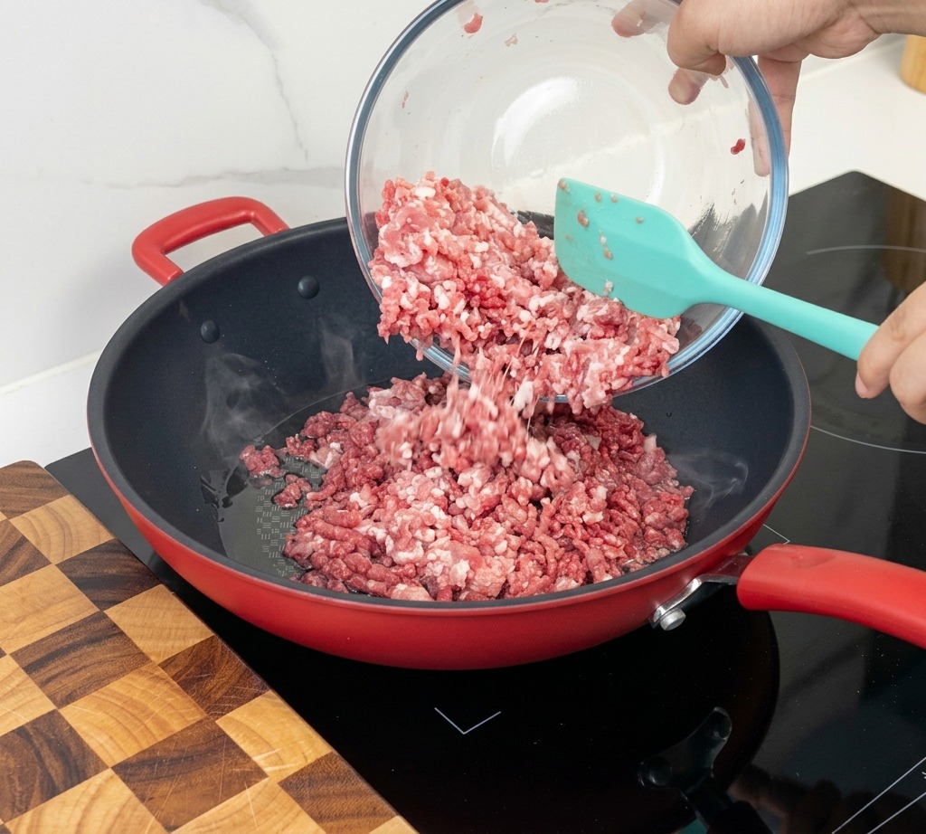 Carne macinata cruda versata da una ciotola di vetro in una padella rossa ben calda usando una spatola in silicone.