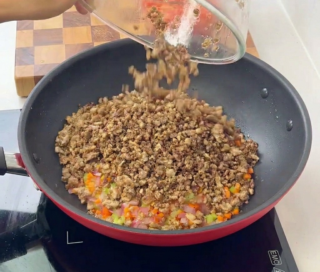 La carne macinata cotta che viene riversata dalla ciotola di vetro nella padella con il soffritto di verdure.