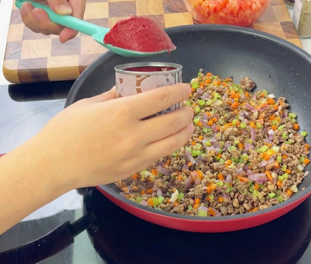 Una spatola azzurra con una porzione di concentrato di pomodoro rosso scuro sopra la padella con carne e verdure.