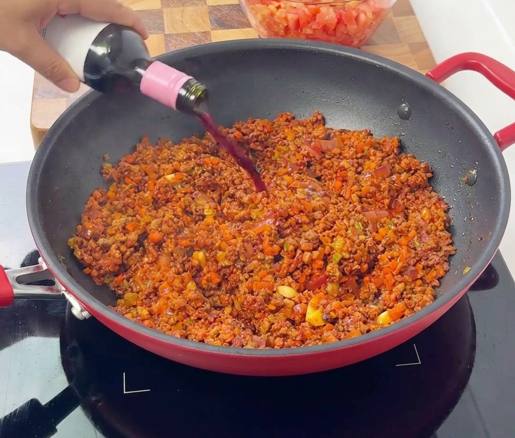Vino rosso versato da una bottiglia in una padella rossa con gli ingredienti del ragù.