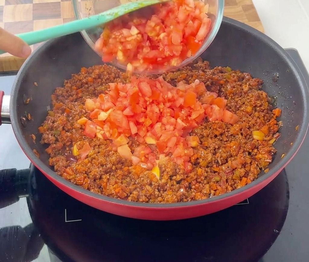 Polpa di pomodoro versata da una ciotola di vetro trasparente nella padella con il ragù in cottura.