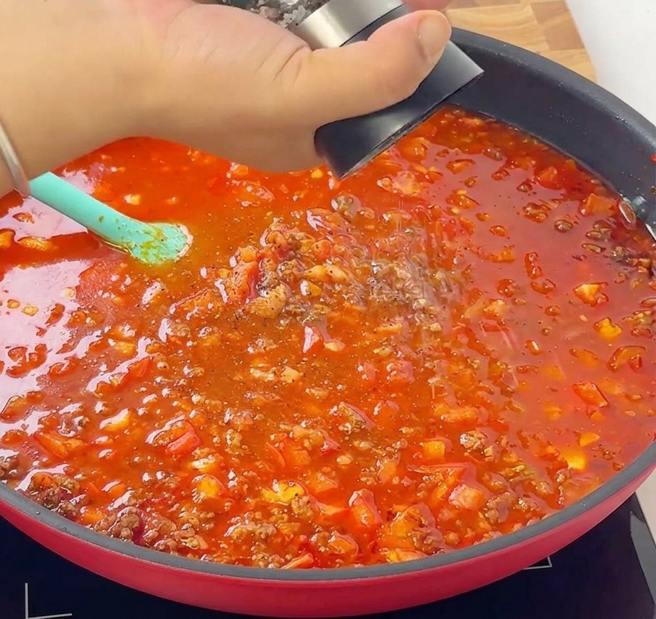 Una mano che utilizza un macinapepe per aggiungere pepe nero fresco al ragù che sobbolle in padella.