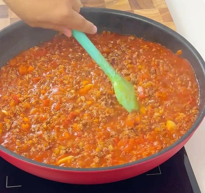 Una spatola verde chiaro che mescola un ragù alla bolognese denso, ricco e ben cotto in una padella rossa.