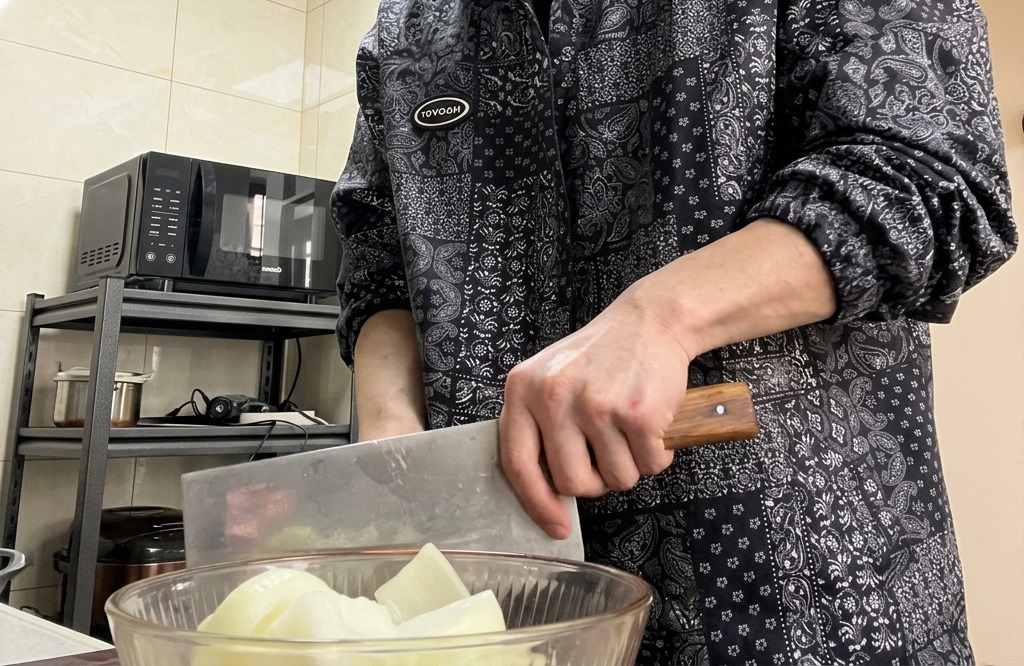 Mani che usano un grosso coltello da cucina per tritare una cipolla su un tagliere di legno.