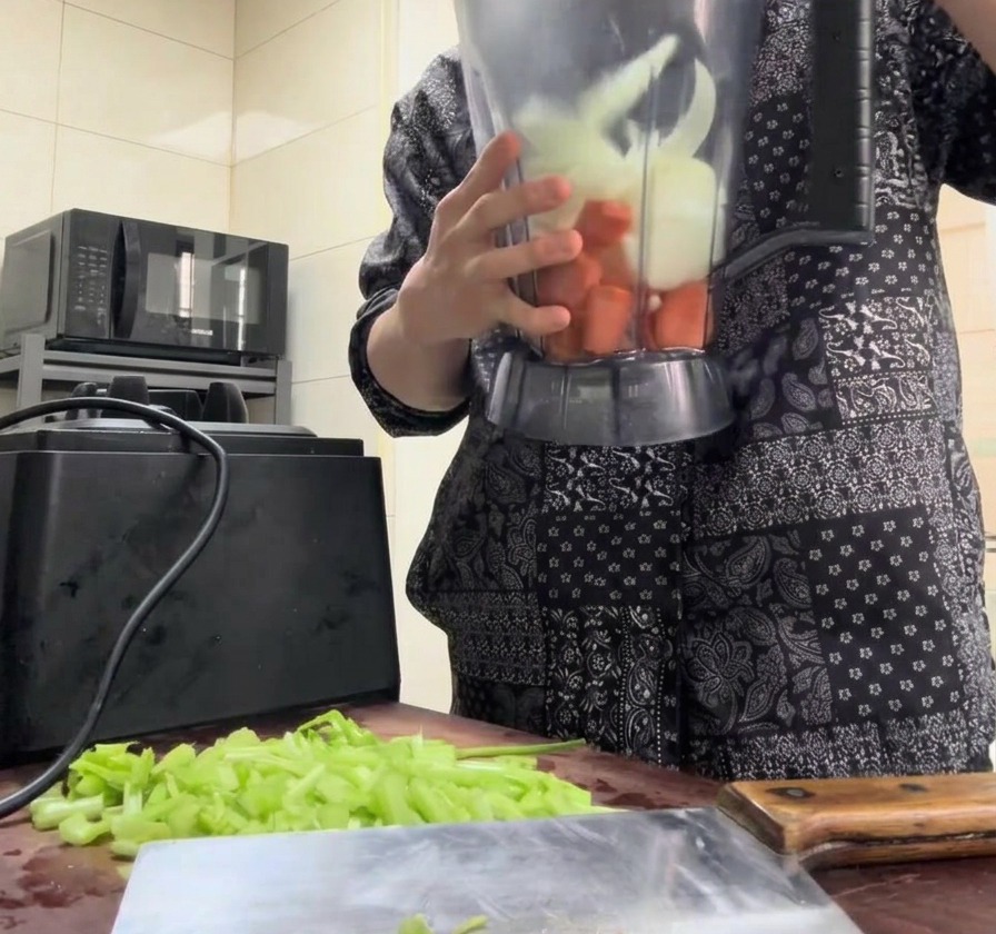 Carote, cipolle e sedano a pezzi versati nel boccale di un frullatore trasparente.