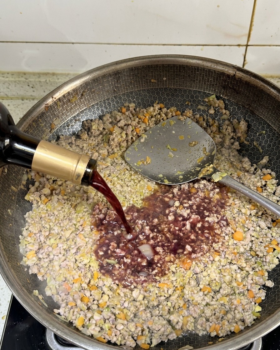 Vino rosso scuro versato da una bottiglia nella padella con la carne macinata rosolata.