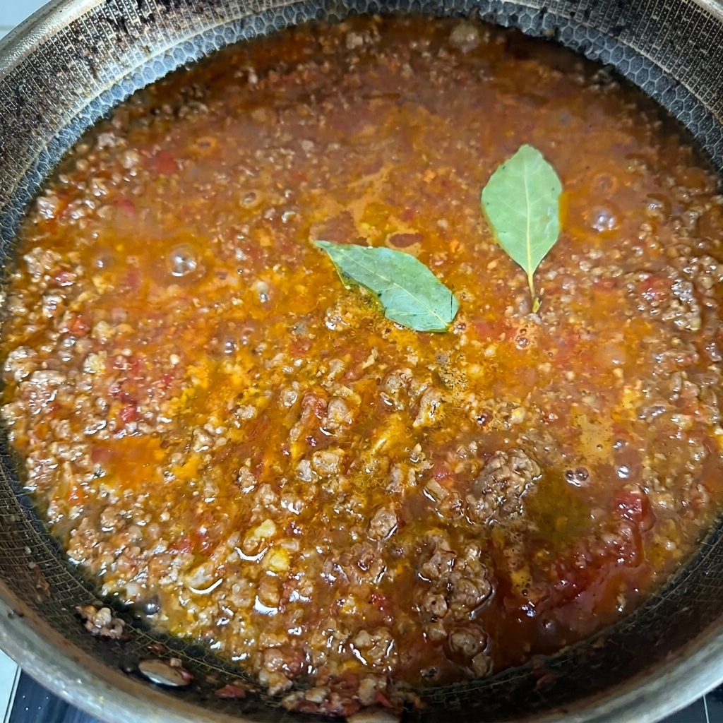 Primo piano di un sugo di carne ricco e denso che sobbolle in una grande pentola con due foglie di alloro verde in superficie.