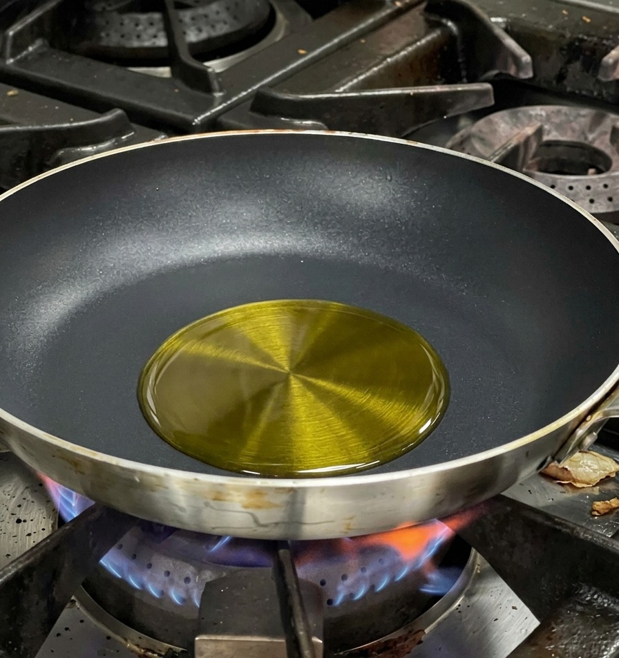 Olio doliva che brilla al centro di una padella antiaderente nera su una fiamma a gas blu.
