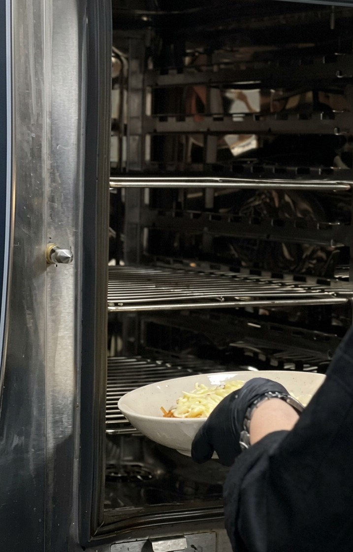 Un cuoco con guanti neri inserisce una ciotola bianca di pasta ricoperta di formaggio in un grande forno industriale in acciaio inossidabile.