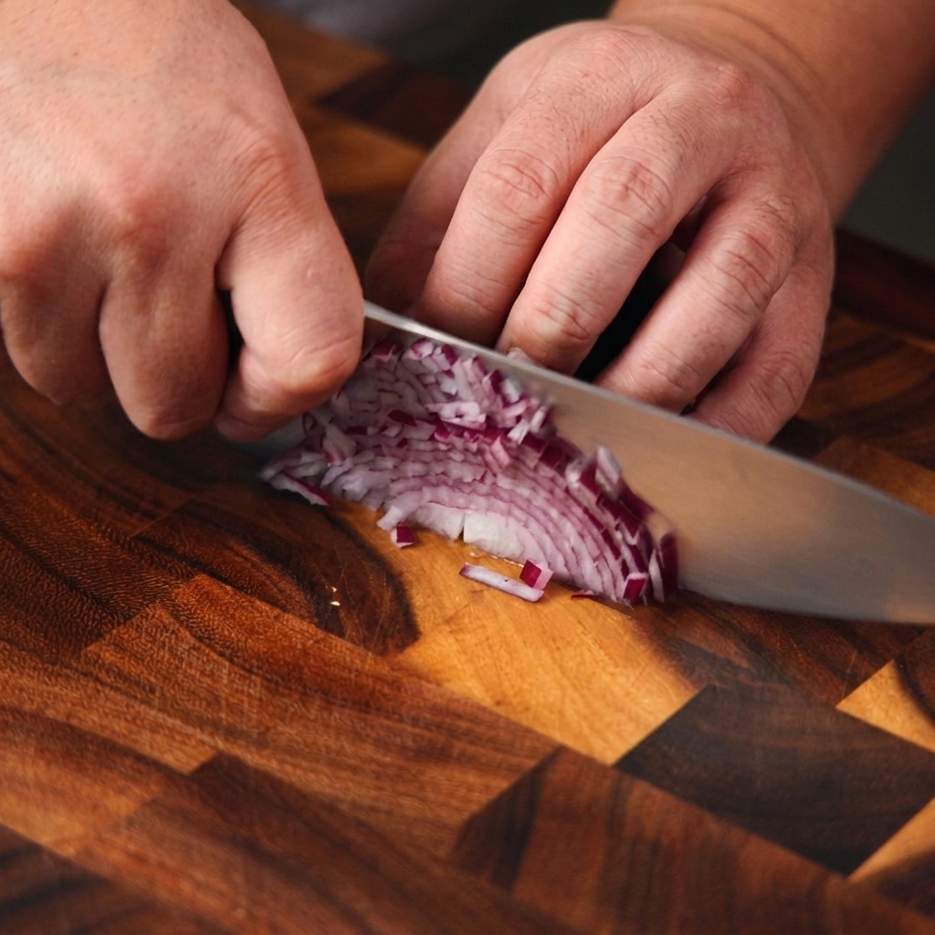 Primo piano di mani che usano un coltello da chef per tritare finemente una cipolla viola su un tagliere di legno.