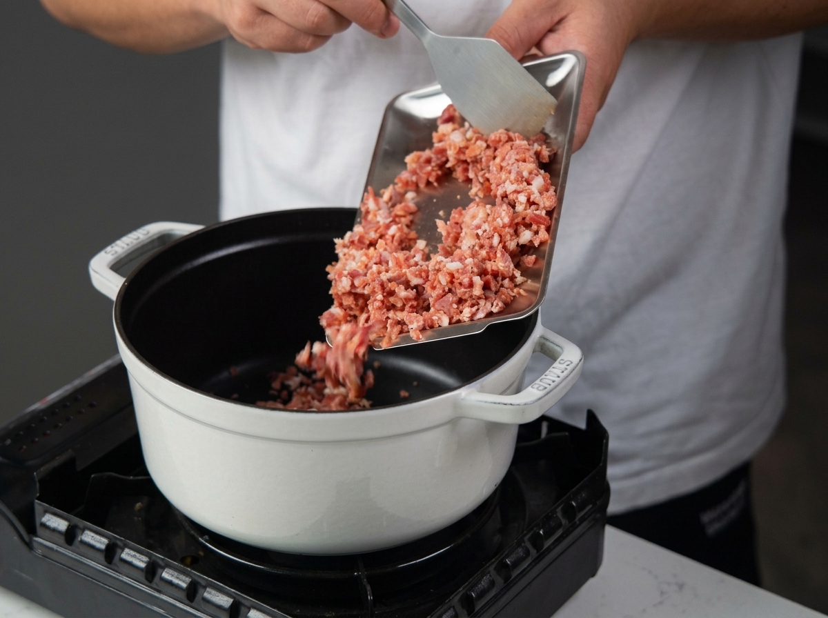 Pancetta a cubetti versata da un vassoio di metallo in una pentola di ghisa smaltata bianca su un fornello portatile.