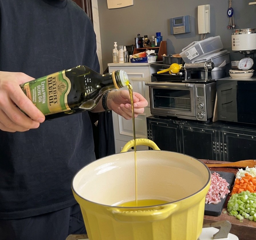 Olio doliva che viene versato da una bottiglia verde in una pentola gialla calda, con la pancetta a cubetti appoggiata su un tagliere lì vicino.