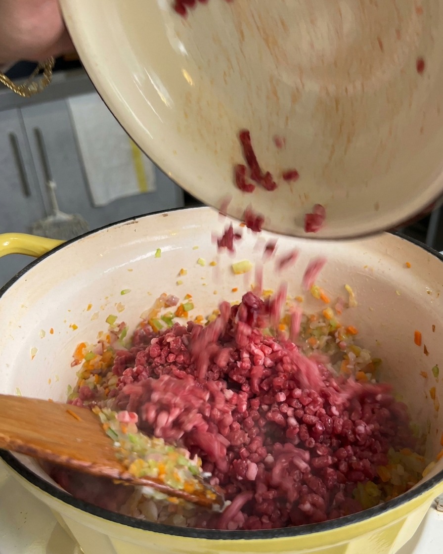 La carne macinata viene versata da una ciotola in una pentola contenente il soffritto di carote, sedano e cipolle.