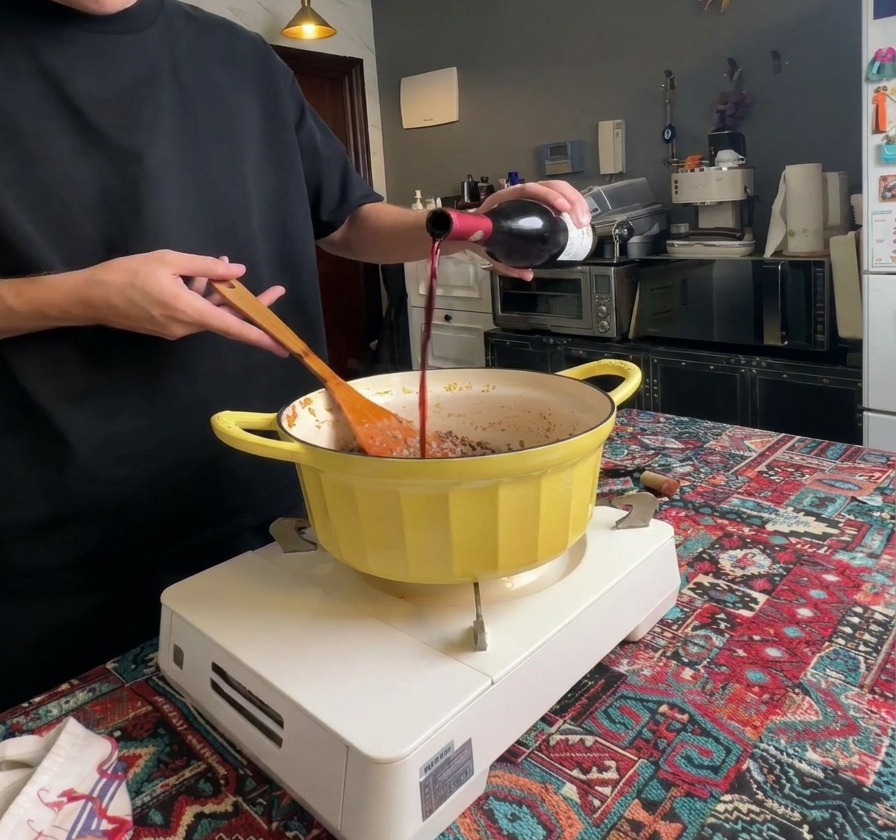 Vino rosso versato da una bottiglia di vetro scuro in una pentola contenente la carne macinata ben rosolata.
