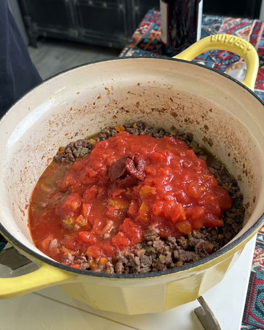 Un cucchiaio di concentrato di pomodoro denso e scuro adagiato sopra la passata e la carne macinata in una pentola gialla.