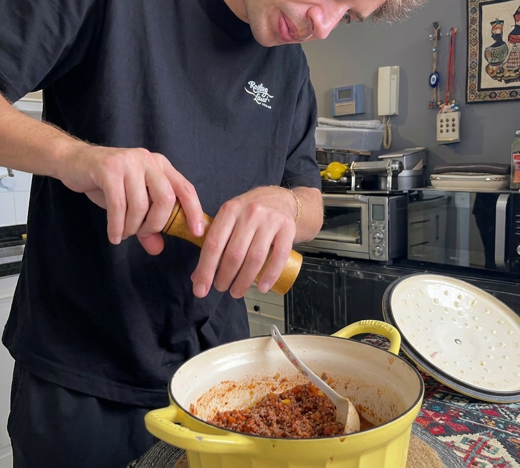 Una persona usa un macinapepe in legno per aggiungere pepe nero fresco al denso ragù di carne allinterno di una casseruola gialla.