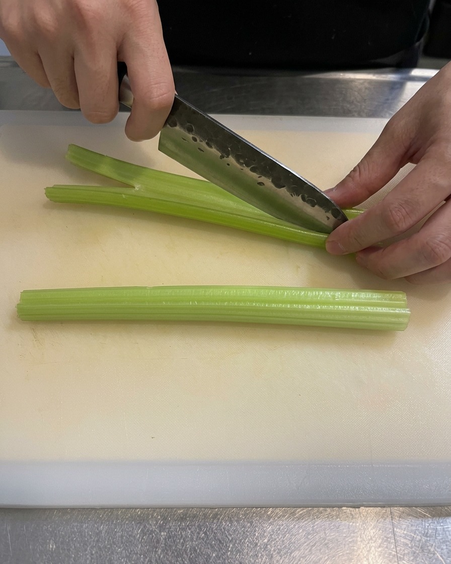 Un primo piano delle mani di uno chef che taglia a piccoli pezzi coste di sedano verde su un tagliere bianco pulito con un coltello affilato.