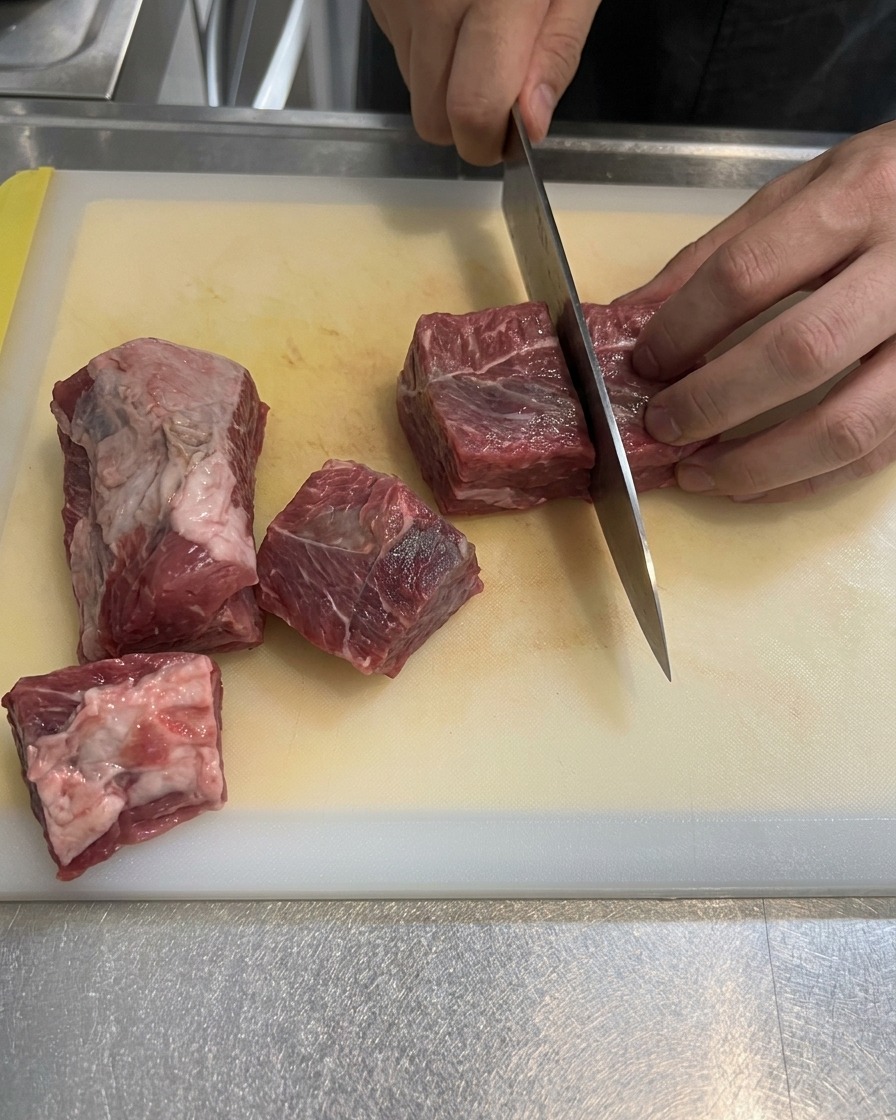 Punta di petto di manzo cruda e venata di grasso che viene tagliata a grossi cubi su un tagliere bianco.