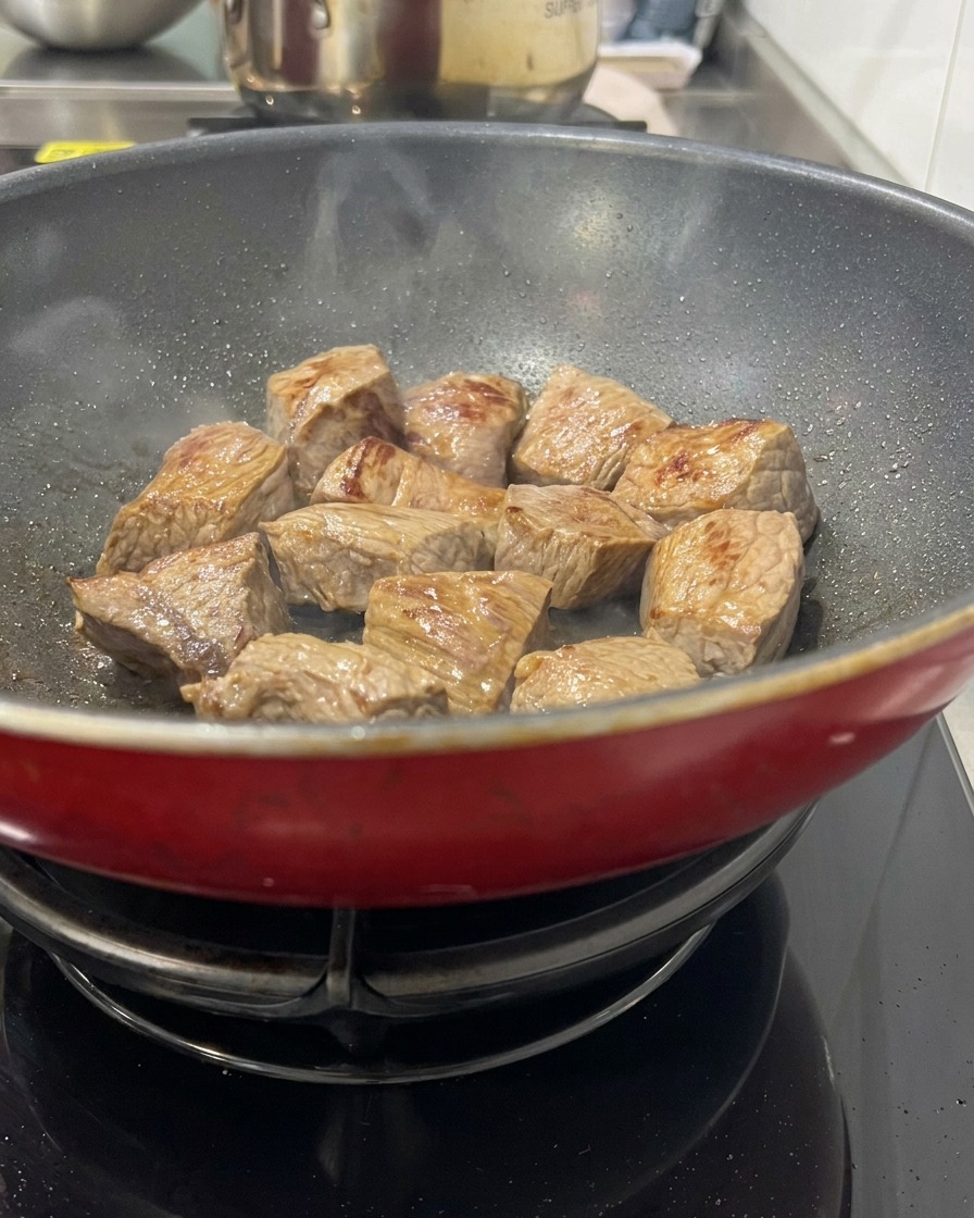 Cubi di carne di manzo che rosolano in una pentola rossa calda, mostrando superfici dorate e un leggero vapore che sale.