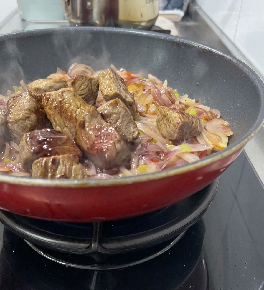 Cubi di carne rosolata adagiati su un letto di cipolle e sedano in una pentola rossa, con laggiunta di vino per sfumare.